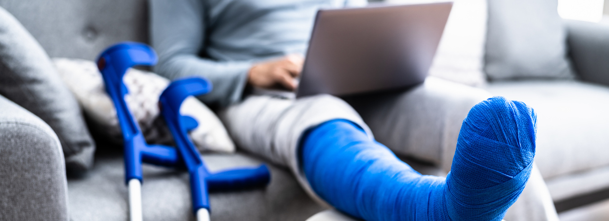 An injured person sitting on the couch working on a laptop with a broken leg in a cast