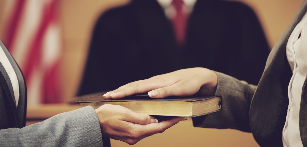 Lawyer holding a bible and witness hand over bible while judge in background.